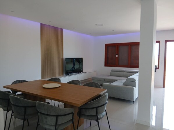 A modern living room with a wooden dining table, grey chairs, a grey sofa, and a television on a white cabinet.