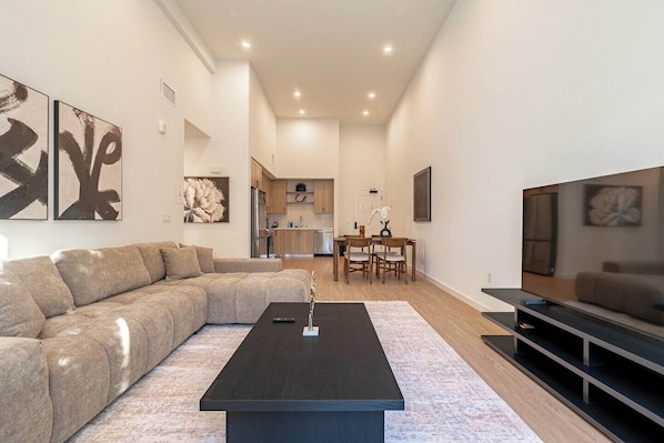 A modern living room with a sectional sofa, a coffee table, and a TV. There is a dining area with a table and chairs, and a kitchen in the background.