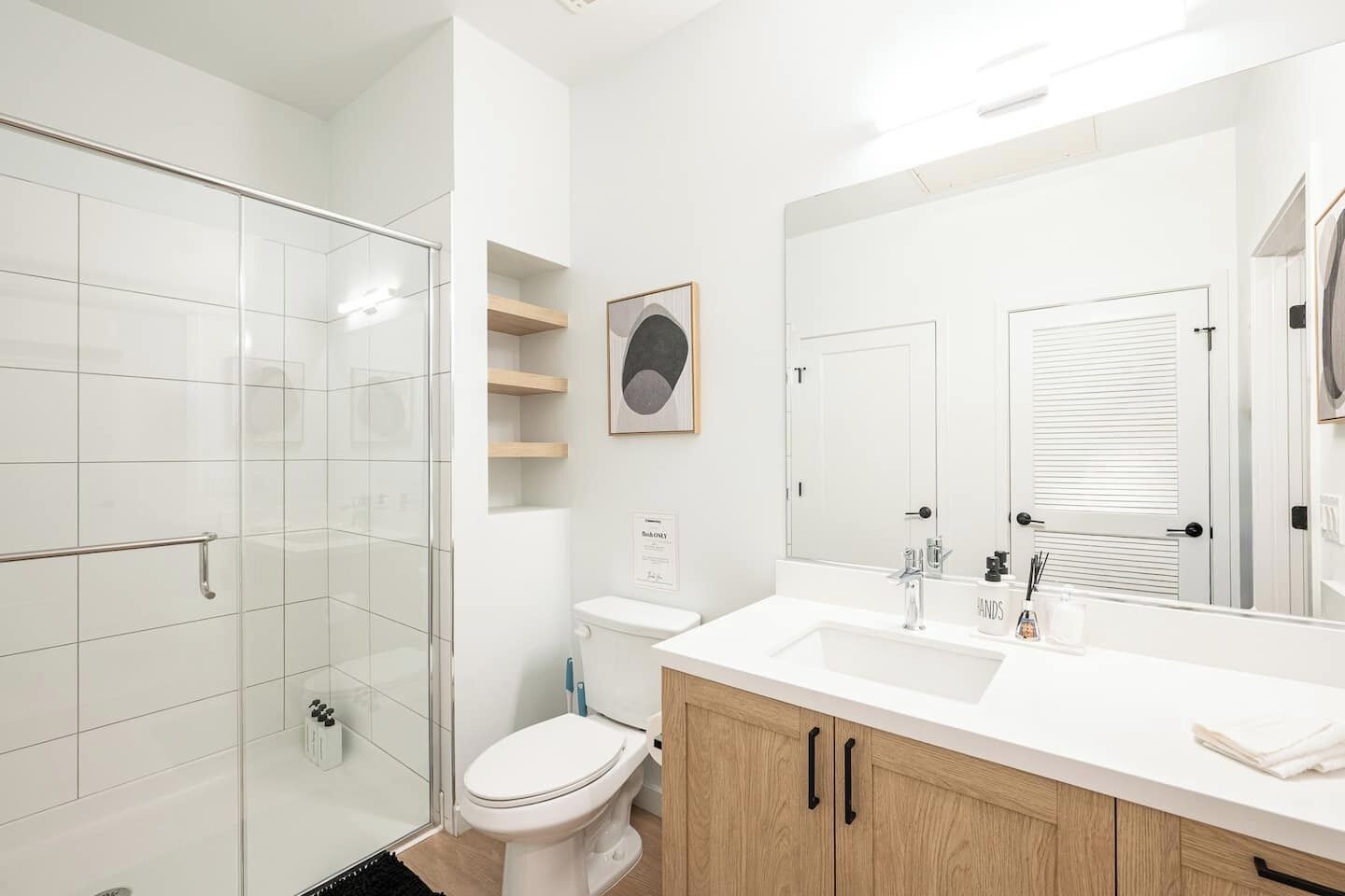 A modern bathroom with a glass shower enclosure, a white toilet, and a wooden vanity with a large mirror.