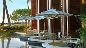 A poolside area with multiple tables and chairs, surrounded by palm trees and a building with wooden shutters. - Experience Island Paradise at The Grand Islander by Hilton - 2 Bd. Ocean View (Honolulu)