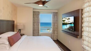 A hotel room with a bed, a TV mounted on the wall, a ceiling fan, and a window with a view of the ocean. - Experience Island Paradise at The Grand Islander by Hilton - 2 Bd. Ocean View (Honolulu)