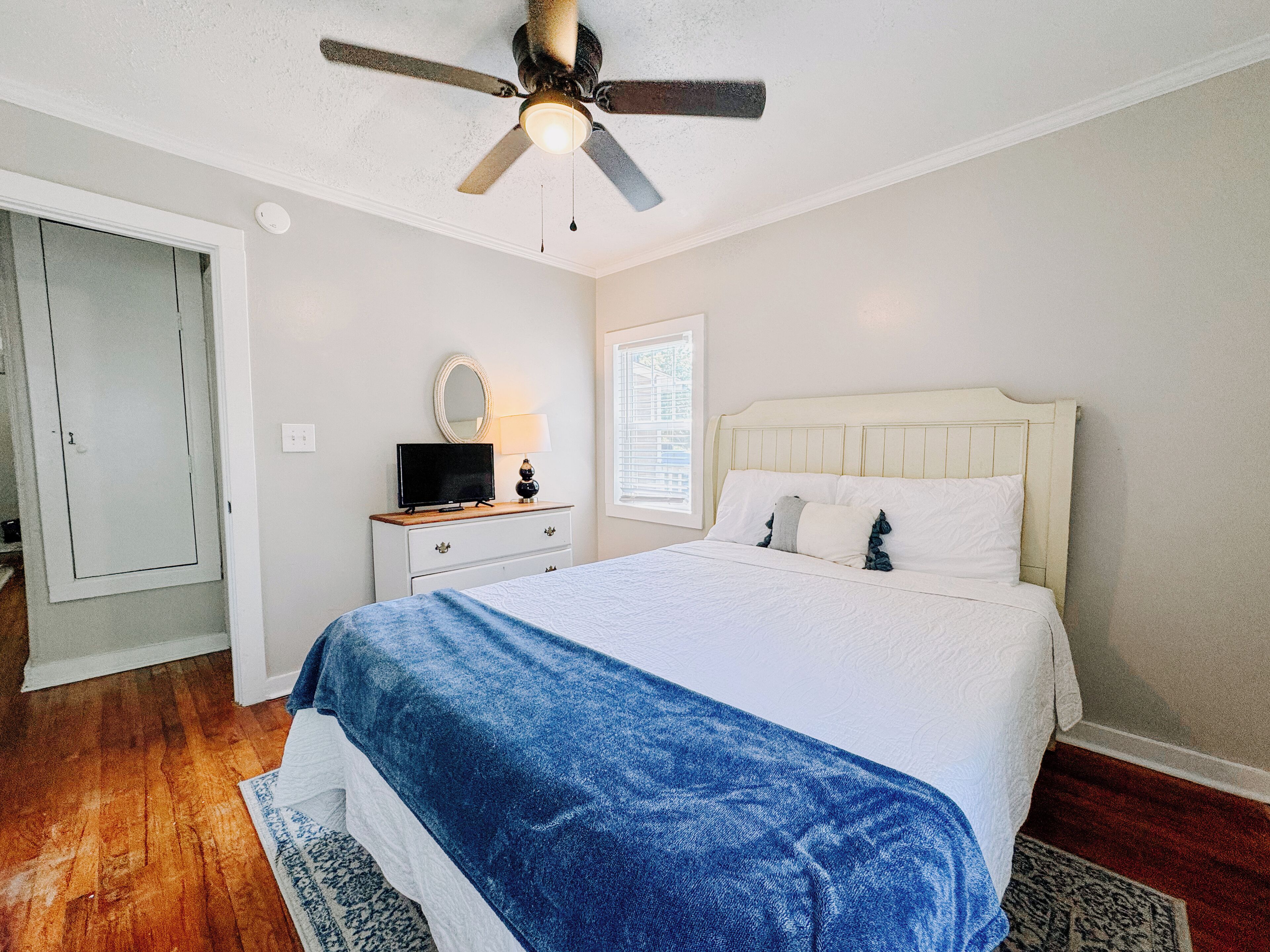 A bedroom with a bed, a ceiling fan, a nightstand with a lamp, a dresser, and a window with blinds.