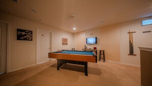 A pool table in a room with beige walls, a door, and a television mounted on the wall. - Go 13 Windsor AT Westside (Kissimmee)