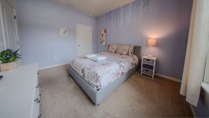 A bedroom with a bed, a nightstand, a lamp, a door, and a wall with a floral pattern. - Go 13 Windsor AT Westside (Kissimmee)