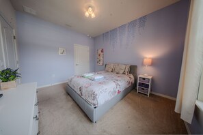 A bedroom with a bed, a nightstand, a lamp, a door, and a wall with a floral pattern. - Go 13 Windsor AT Westside (Kissimmee)