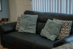 A black leather sofa with two patterned cushions, a small table with a bowl, and a wall with light switches. - Go 13 Windsor AT Westside (Kissimmee)