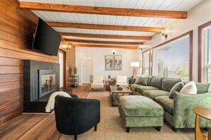 Ein modernes Wohnzimmer mit Kamin, Holzbalken, einem Fernseher und einem großen Fenster.