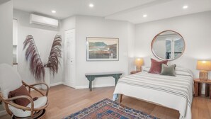 A bedroom with a bed, bedside lamps, a bench, a chair, a mirror, and a wall-mounted air conditioner.