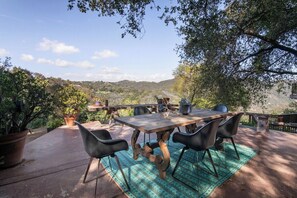 An outdoor dining area with a table and chairs, surrounded by potted plants and overlooking a hilly landscape.