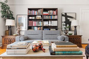 Ein Wohnzimmer mit grauem Sofa, einem Couchtisch mit Büchern und einem Globus, einem Bücherregal voller Bücher und zwei Lampen.