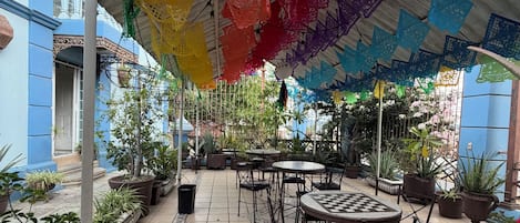 Un espace extérieur aménagé en terrasse, avec des tables et des chaises, entouré de plantes en pot et surmonté d’un auvent aux couleurs vives.