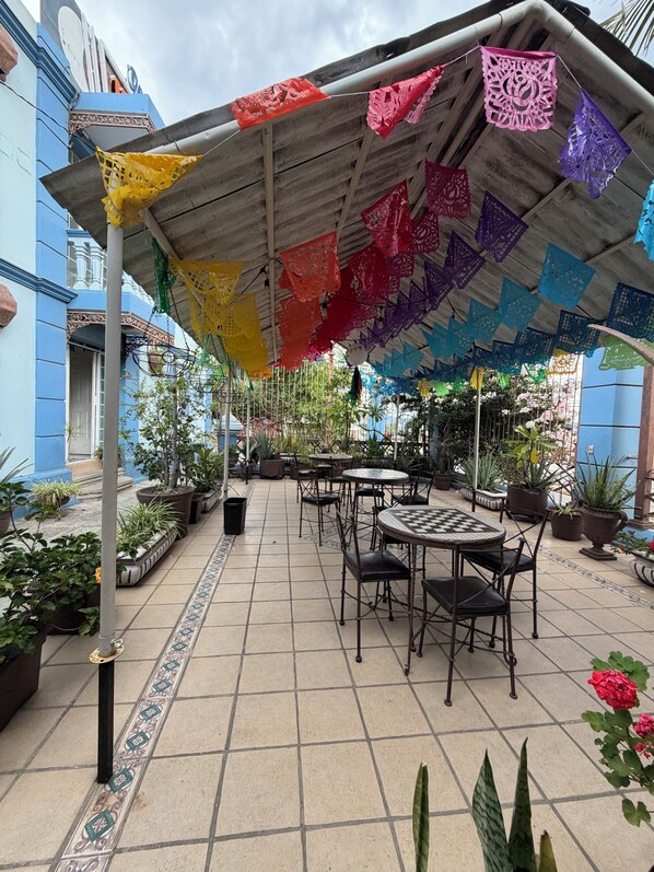 An outdoor patio area with tables and chairs, surrounded by potted plants and a colorful canopy.