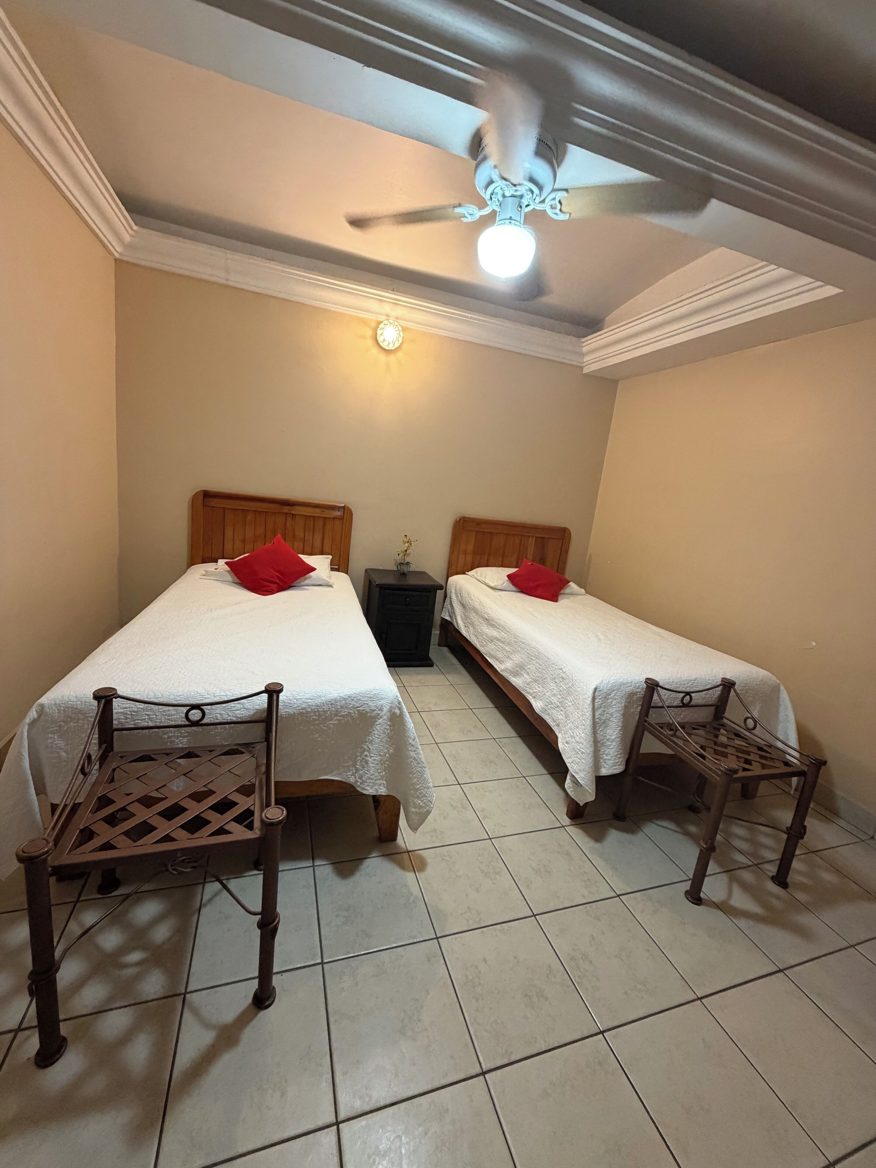 Una habitación con dos camas, cada una con cabecera de madera y almohada roja, un ventilador de techo y una mesita entre las camas.
