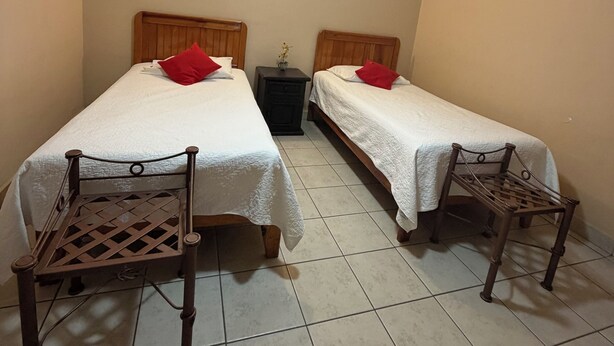 Ein Zimmer mit zwei Betten, jeweils mit einem Holz-Kopfstück und einem roten Kissen, einem Deckenventilator und einem kleinen Tisch zwischen den Betten.