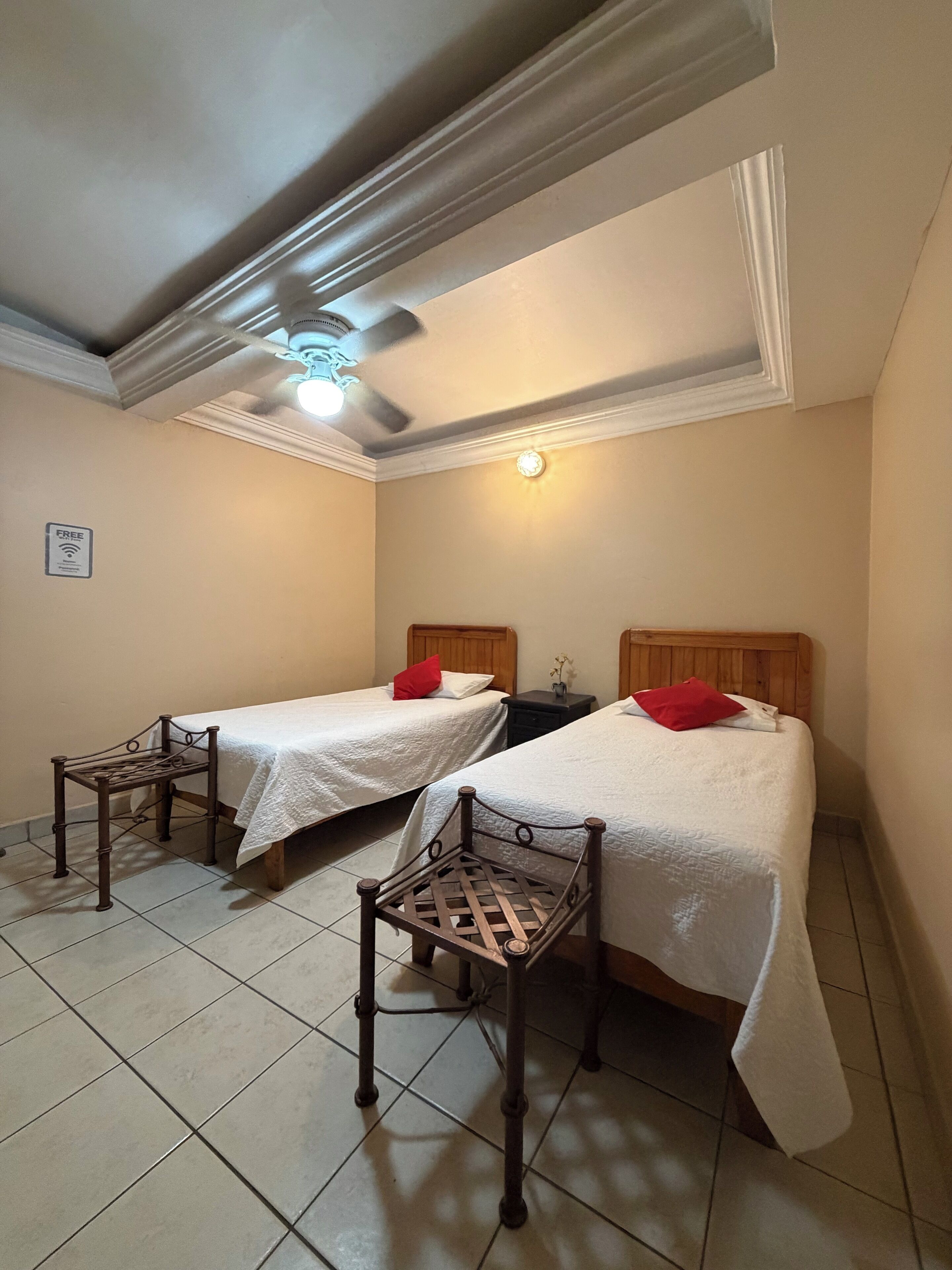 Quarto de hotel com duas camas, cada uma com cabeceira de madeira e travesseiro vermelho, uma mesinha e um ventilador de teto.