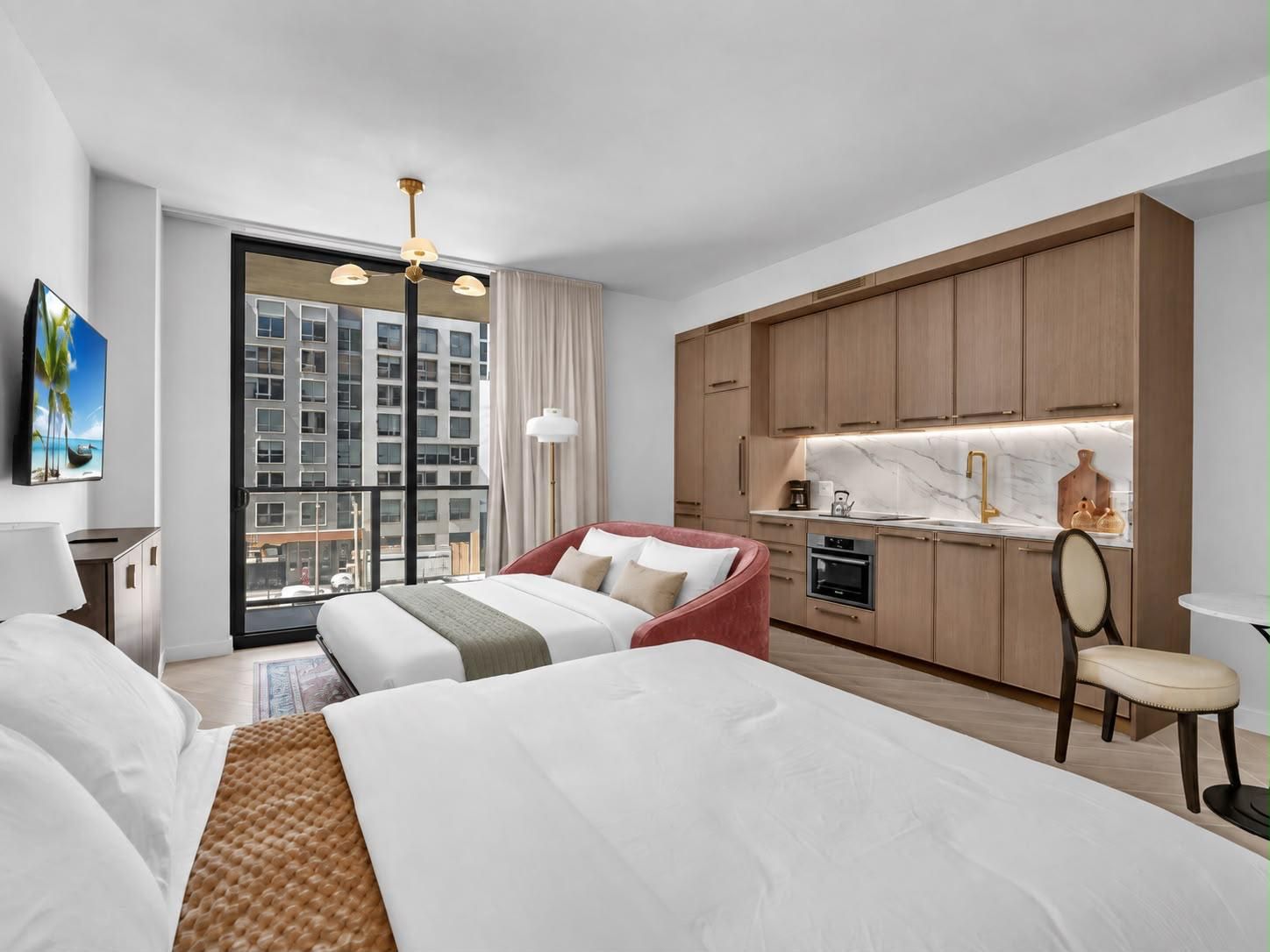 A hotel room with a bed, a desk, a chair, a kitchenette, and a large window with a view of a building.