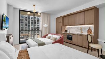 A hotel room with a bed, a desk, a chair, a kitchenette, and a large window with a view of a building.