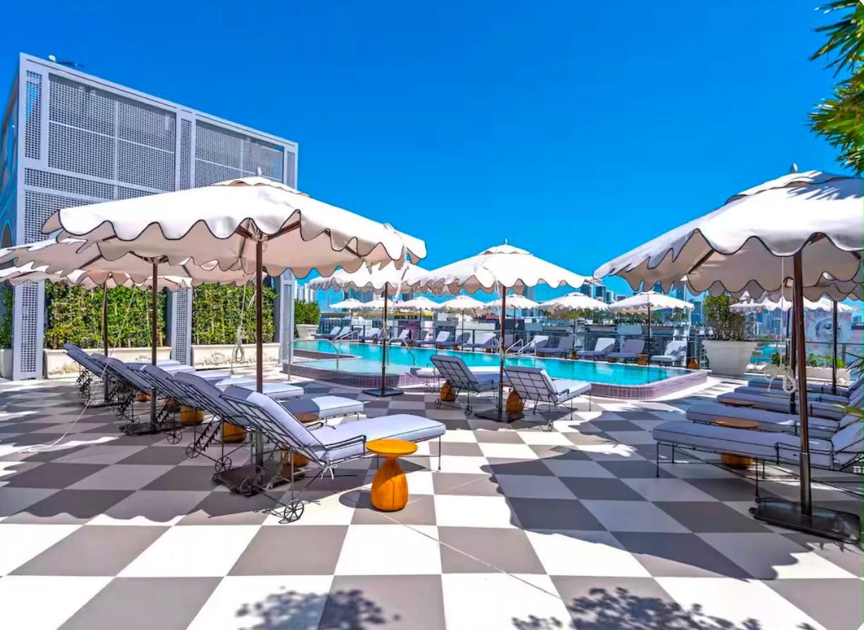 Un espace aménagé au bord de la piscine, avec des chaises longues et des parasols, un sol à carreaux et un ciel d’un bleu limpide.