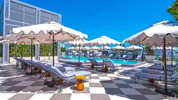 Un espace aménagé au bord de la piscine, avec des chaises longues et des parasols, un sol à carreaux et un ciel d’un bleu limpide.