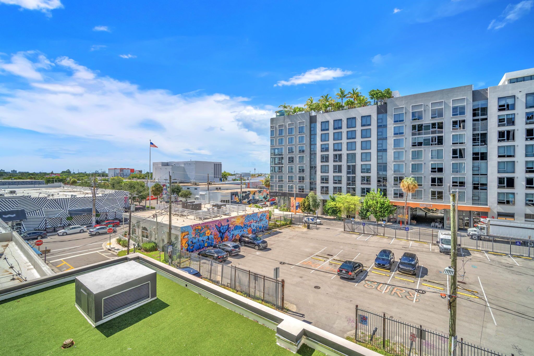 A parking lot with multiple cars, a mural, and modern buildings in the background.