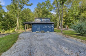 A blue building with a gravel driveway leading to a covered area, surrounded by trees and a grassy landscape. - Waterfront Blue Spruce w Hot Tub & Fire Pit! (Wildwood)