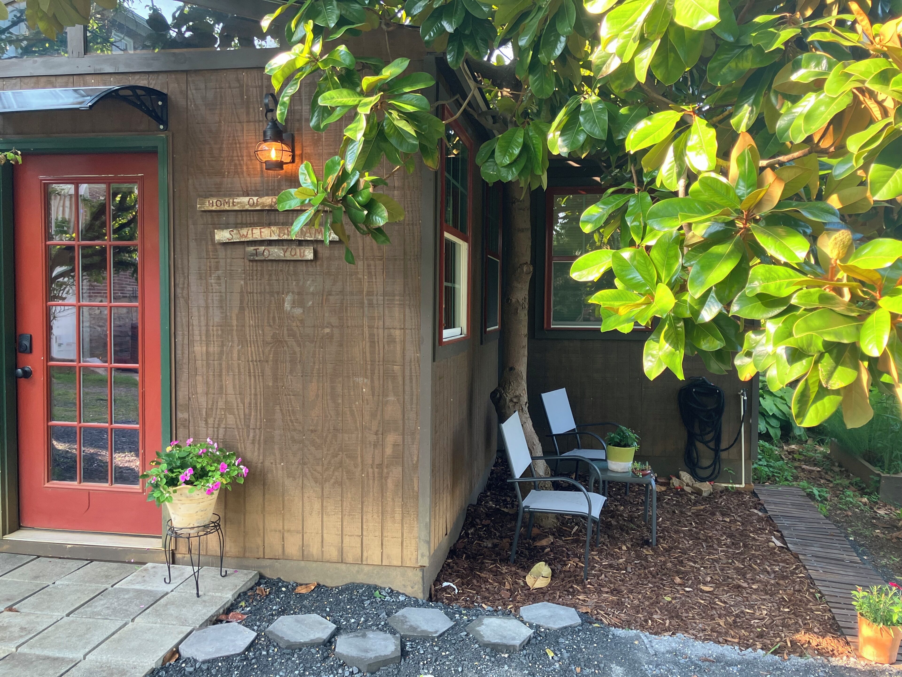 A wooden building with a red door, a sign that reads 'HOME OF SWEETNESS TO YOU,' and a potted plant with pink flowers.