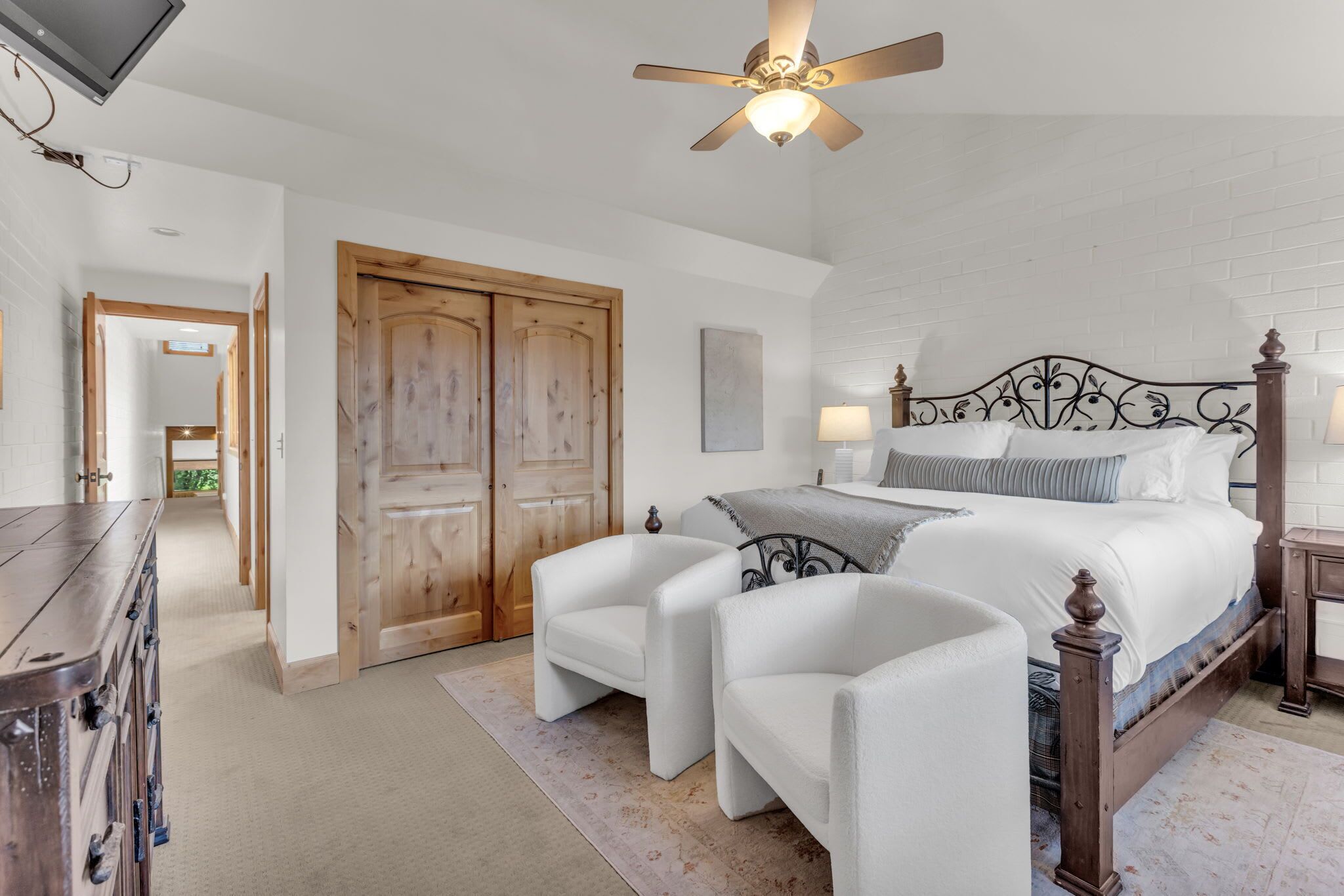 A bedroom with a bed, two chairs, a wooden wardrobe, a ceiling fan, and a TV.