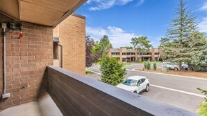 Een balkon met een bakstenen muur, een metalen hekwerk en een helder uitzicht op een parkeerplaats met auto's en een gebouw op de achtergrond.