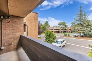 A balcony with a brick wall, a metal railing, and a clear view of a parking lot with cars and a building in the background. - Park City Retreat | A/C + Hot Tub + Shared Pool (Park City)