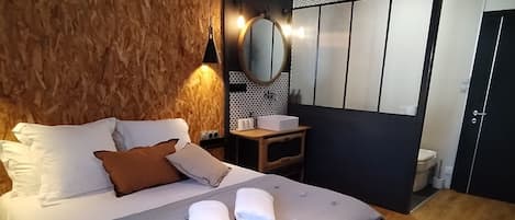 A modern hotel room with a large bed, a wooden headboard, a small bedside table, a mirror, and a view of the outside.