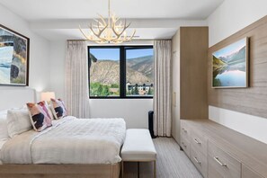 A hotel room with a large bed, a bedside table, a desk, and a view of mountains through a window. - Frontgate | Luxury Condo W/Resort Amenities (Avon)