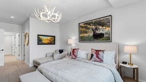 Ein modernes Schlafzimmer mit einem großen Bett, einer Kristallleuchtre, einem Bild eines Elchs und einem Nachttisch mit einer Lampe.