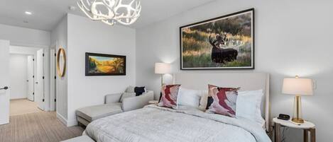 A modern bedroom with a large bed, a chandelier, a painting of a moose, and a bedside table with a lamp.