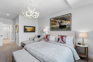 A modern bedroom with a large bed, a chandelier, a painting of a moose, and a bedside table with a lamp. - Frontgate | Luxury Condo W/Resort Amenities (Avon)