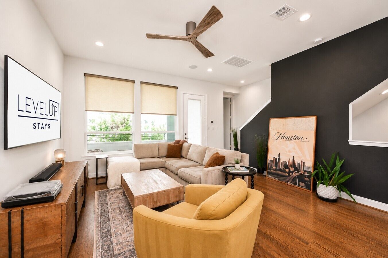 Un salon moderne avec un canapé d’angle, une table basse, un fauteuil jaune, un ventilateur de plafond et une œuvre d’art représentant un paysage urbain.