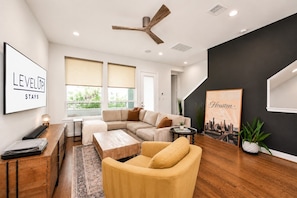 A modern living room with a sectional sofa, a coffee table, a yellow armchair, a ceiling fan, and a cityscape artwork.