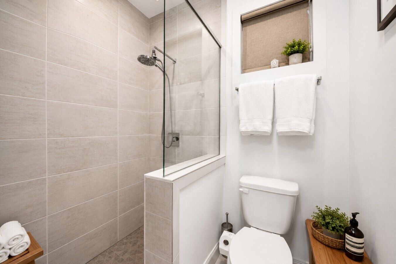 A modern bathroom with a glass-enclosed shower, a toilet, and a small shelf with towels and a plant.
