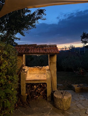 An outdoor cooking area with a stone structure, a fire pit, and a stack of firewood.