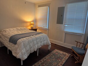 A bedroom with a large bed, a nightstand with a lamp, two windows with blinds, a chair, and a patterned rug.