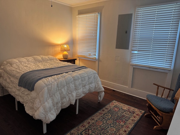 A bedroom with a large bed, a nightstand with a lamp, two windows with blinds, a chair, and a patterned rug.
