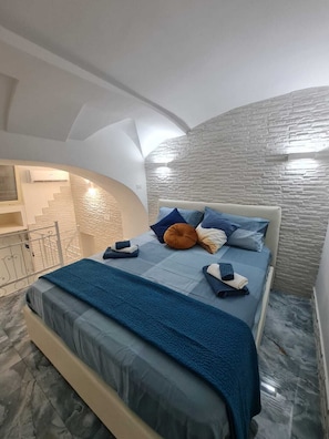 A bedroom with a bed, a blue blanket, and pillows. There is a white railing and a stone wall. - Furnished and fully equipped Piazza Plebiscito House on Via Toledo in downtown Naples (Napoli)