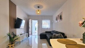 Ein modernes Wohnzimmer mit einer Couch, einem Esstisch mit Platzsets, einem an der Wand befestigten Flachbildfernseher und einer Deckenleuchte.