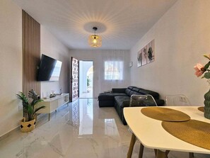 A modern living room with a sofa, a dining table with placemats, a flat-screen TV mounted on the wall, and a ceiling light fixture. - Tranquil oasis in Costa Blanca (VC)