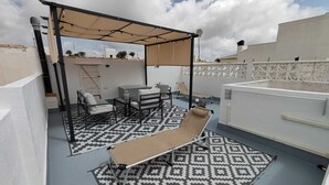 Eine Dachterrasse mit gemustertem Boden, einer Liege, einem Tisch mit Stühlen und einem überdachten Sitzbereich.