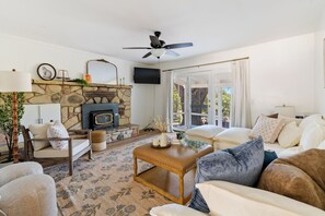 Ein Wohnzimmer mit Kamin aus Stein, Deckenventilator und Blick durch Glastüren.