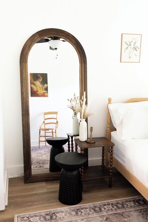 A bedroom with a large mirror, a bed, a chair, a small table with a vase, and a rug.