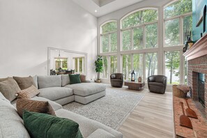 Un salon spacieux avec un grand canapé d’angle, une cheminée et une vue sur les arbres grâce à de grandes fenêtres.