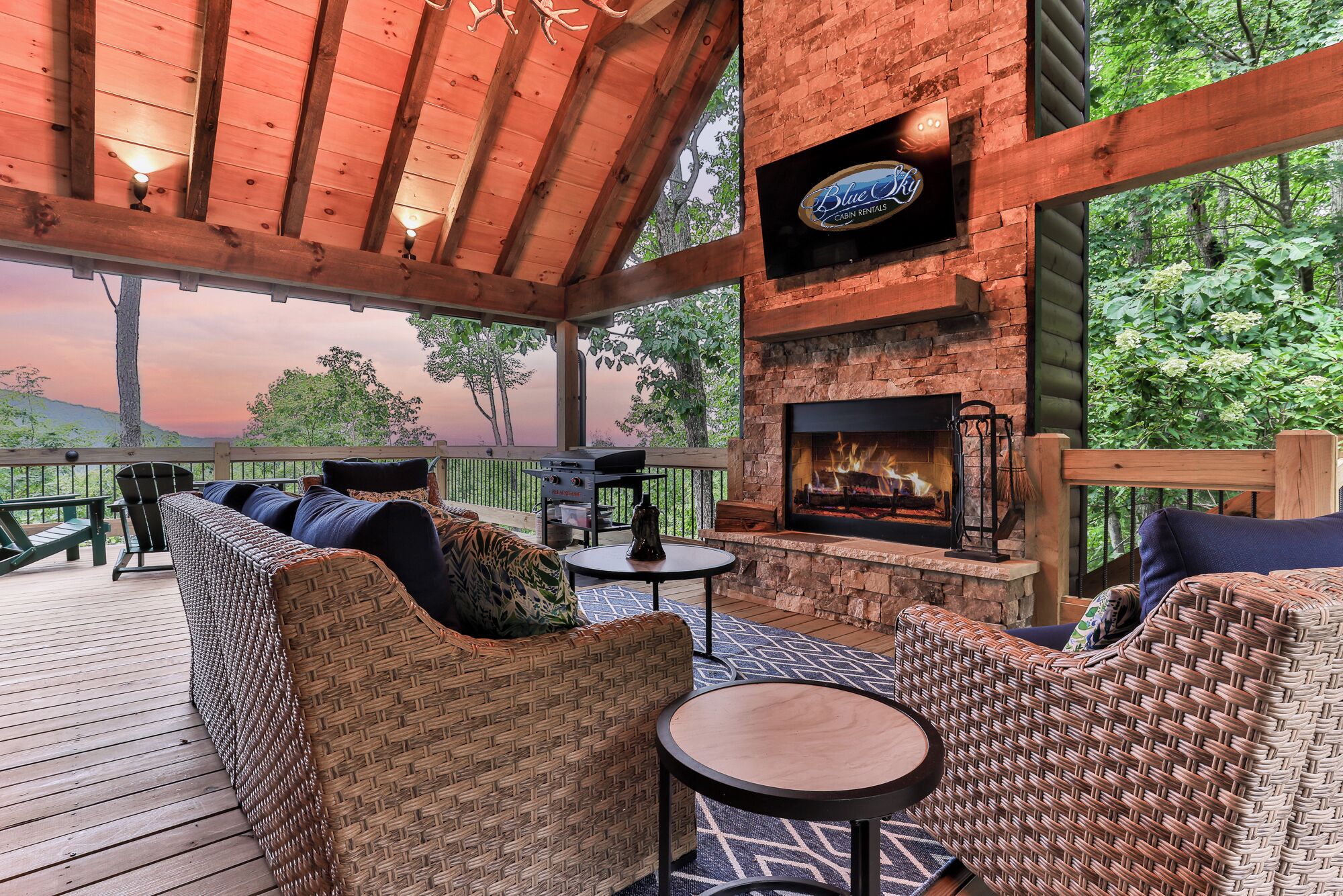 A covered outdoor seating area with wicker furniture, a stone fireplace, and a television screen above it.