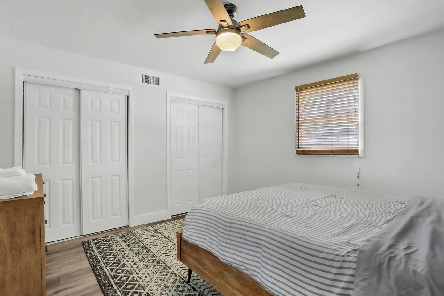 Ein Schlafzimmer mit einem Bett, einem Deckenventilator, einem Fenster mit Jalousien und einem gemusterten Teppich.
