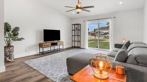 A modern living room with a flat-screen TV, a ceiling fan, a large window with a view, a grey sofa, and a decorative table lamp. - 3BR/2B Retreat - Walk to town (St. Augustine)